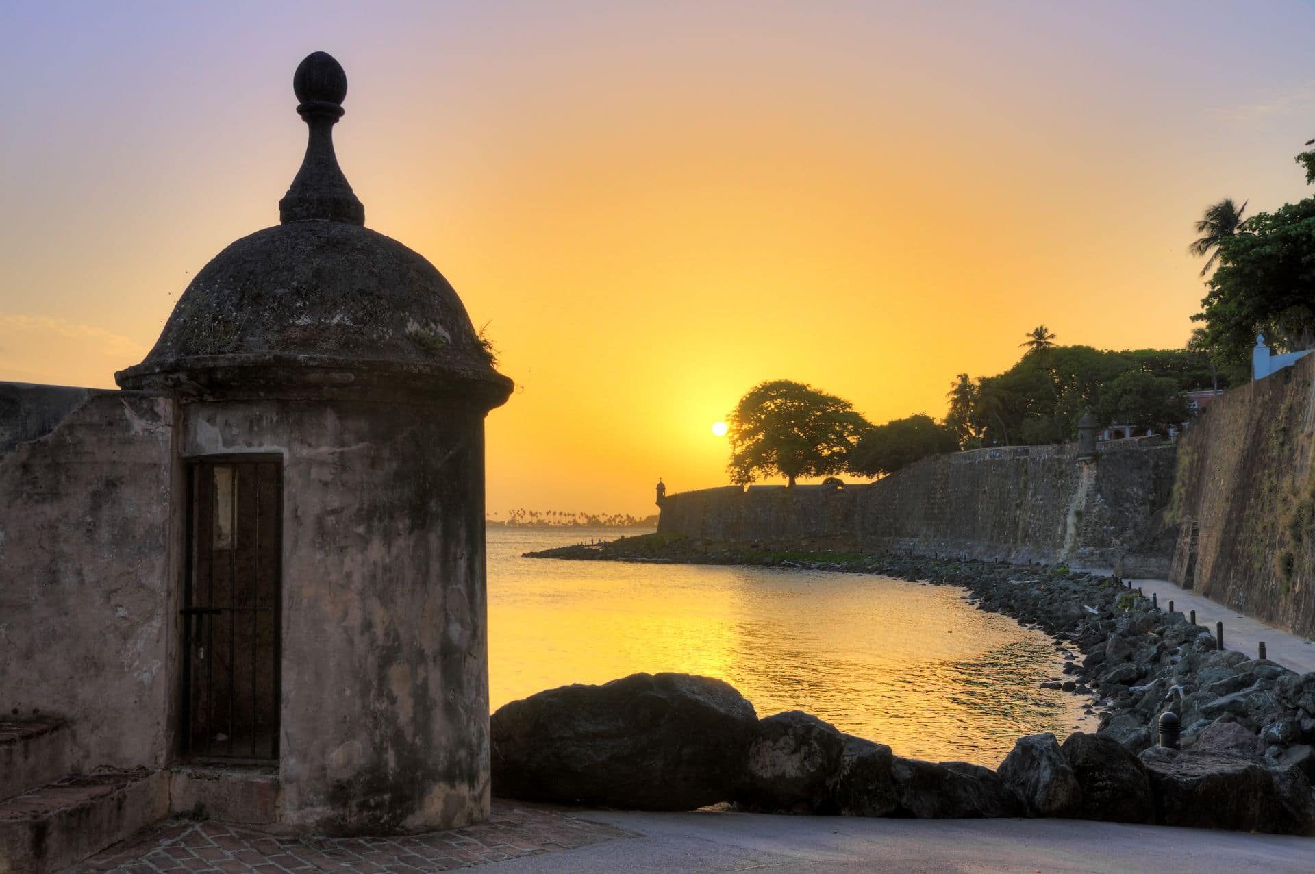 The light from a bright yellow sunset washing over the tower and gray walls of a fort in San Juan Puerto, Rico.