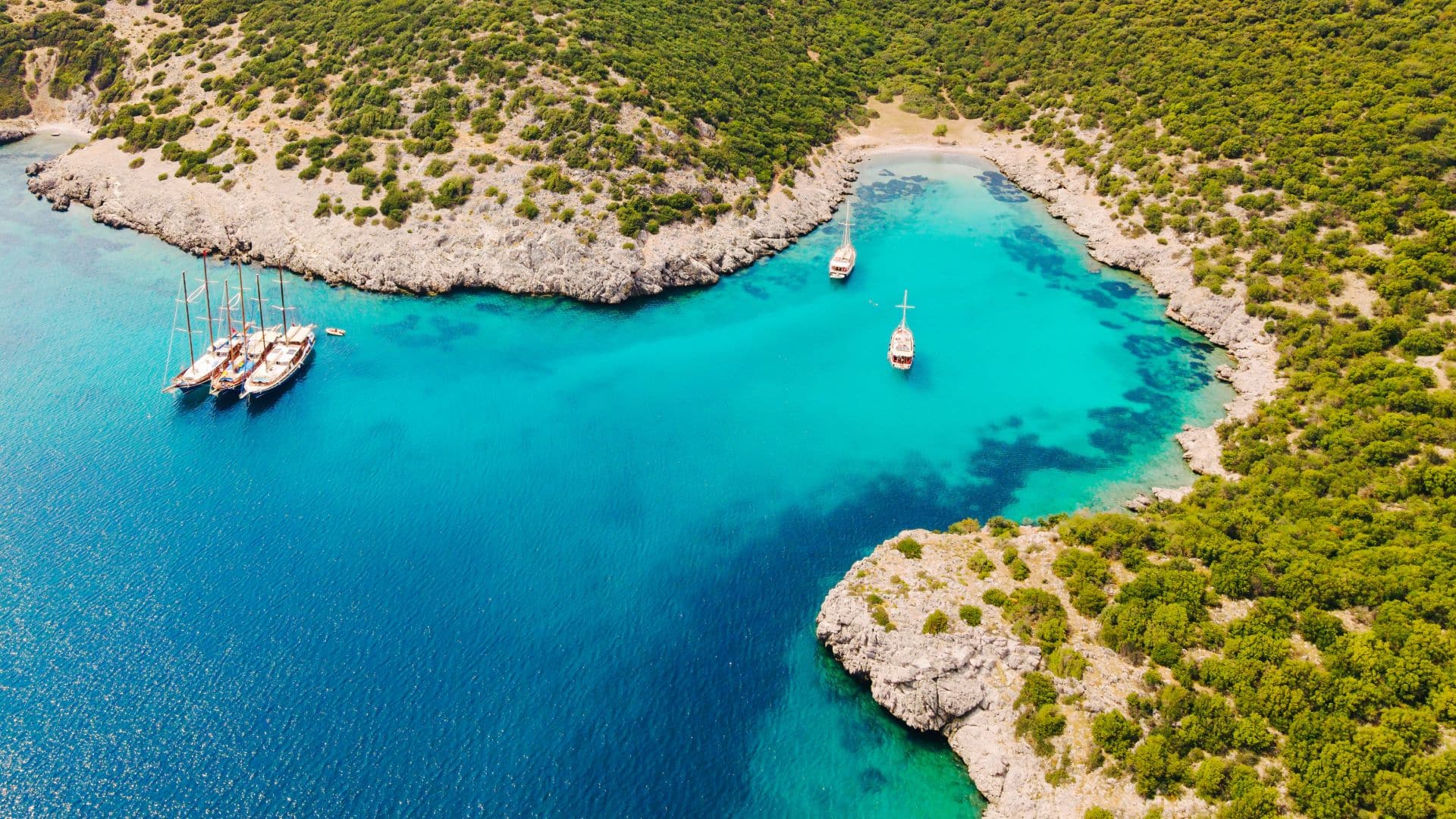 Aerial view of a Mediterranean cove with sailboats, green hills, rocky cliffs, and a secluded sandy beach.