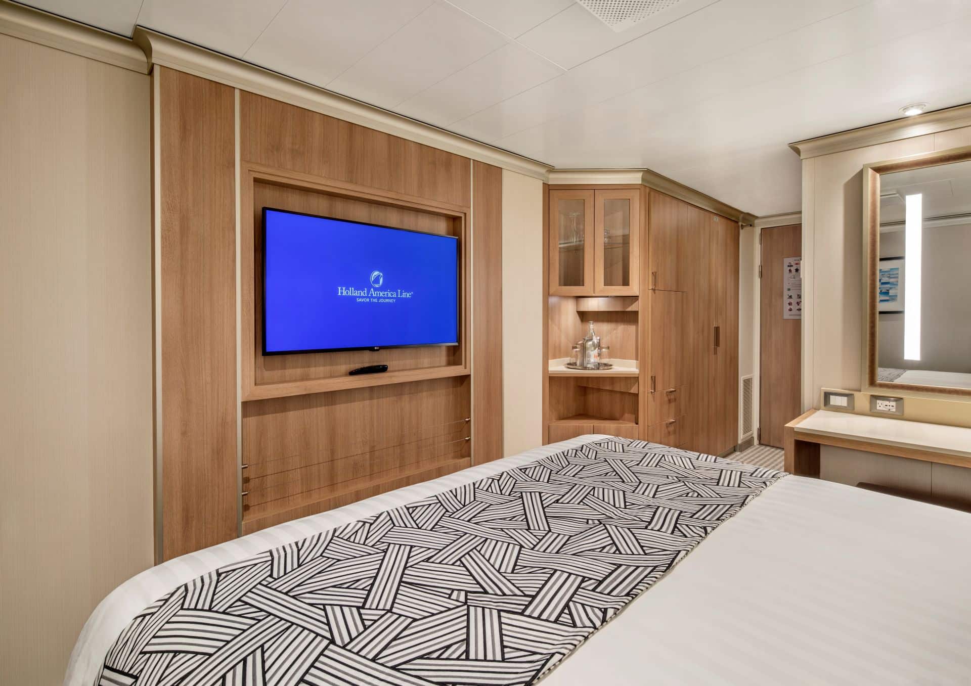 Cruise ship stateroom with bed, wall-mounted TV showing Holland America Line logo, wood cabinetry, and vanity.