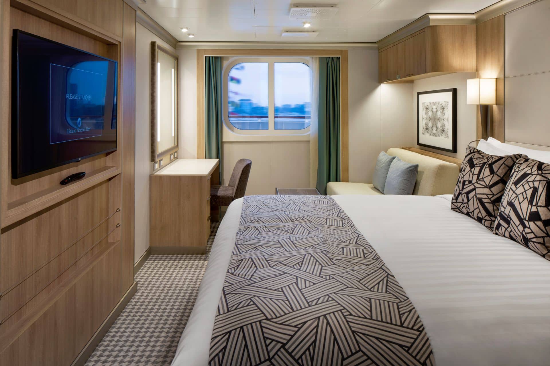 Cruise ship ocean view stateroom with bed, wall mounted TV, desk, window, and wood cabinetry.