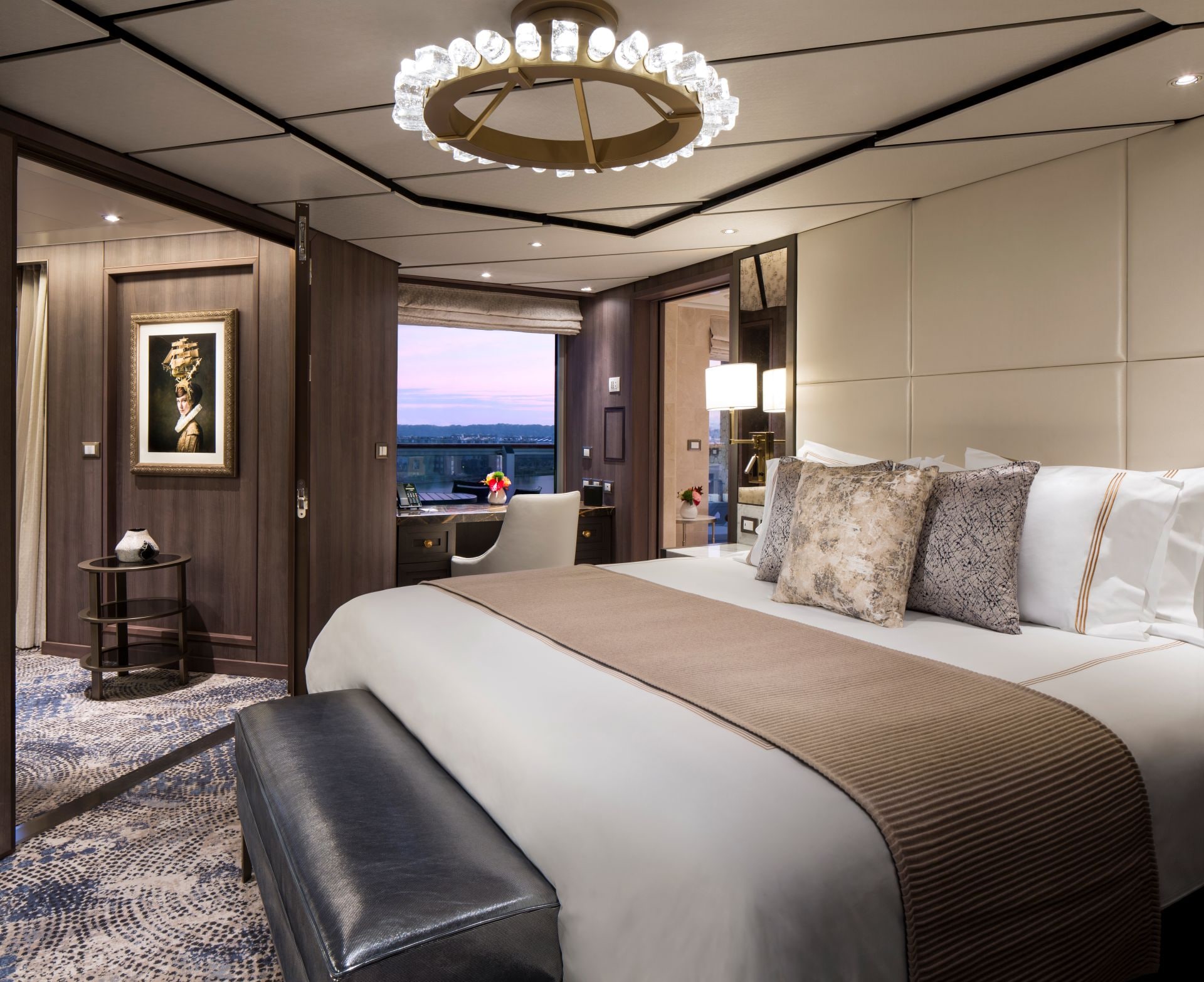 Cruise ship suite with king bed, sitting area, chandelier, desk, and balcony doors.