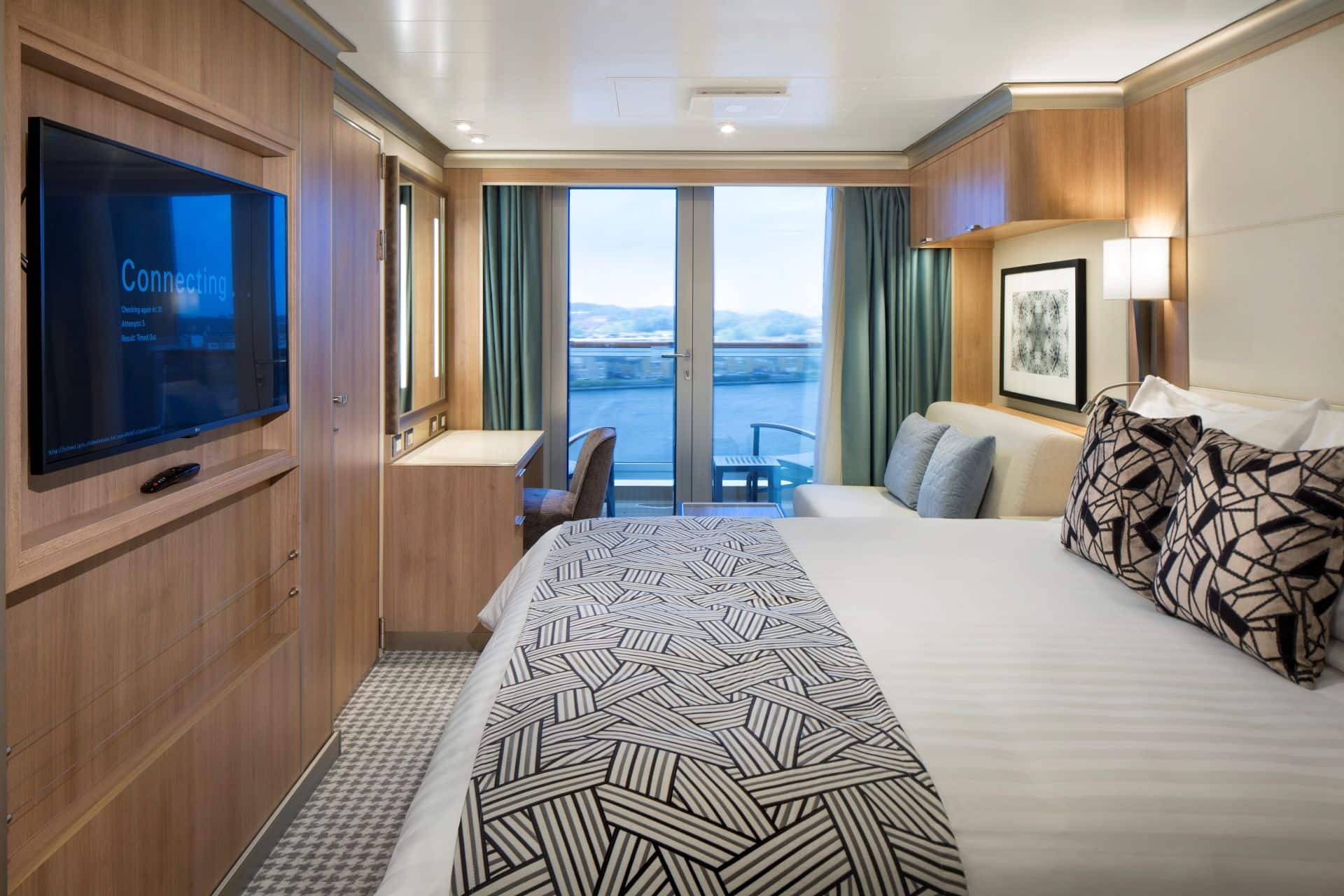 Cruise ship verandah stateroom with bed, wall mounted TV, desk, sliding glass doors, and balcony view.