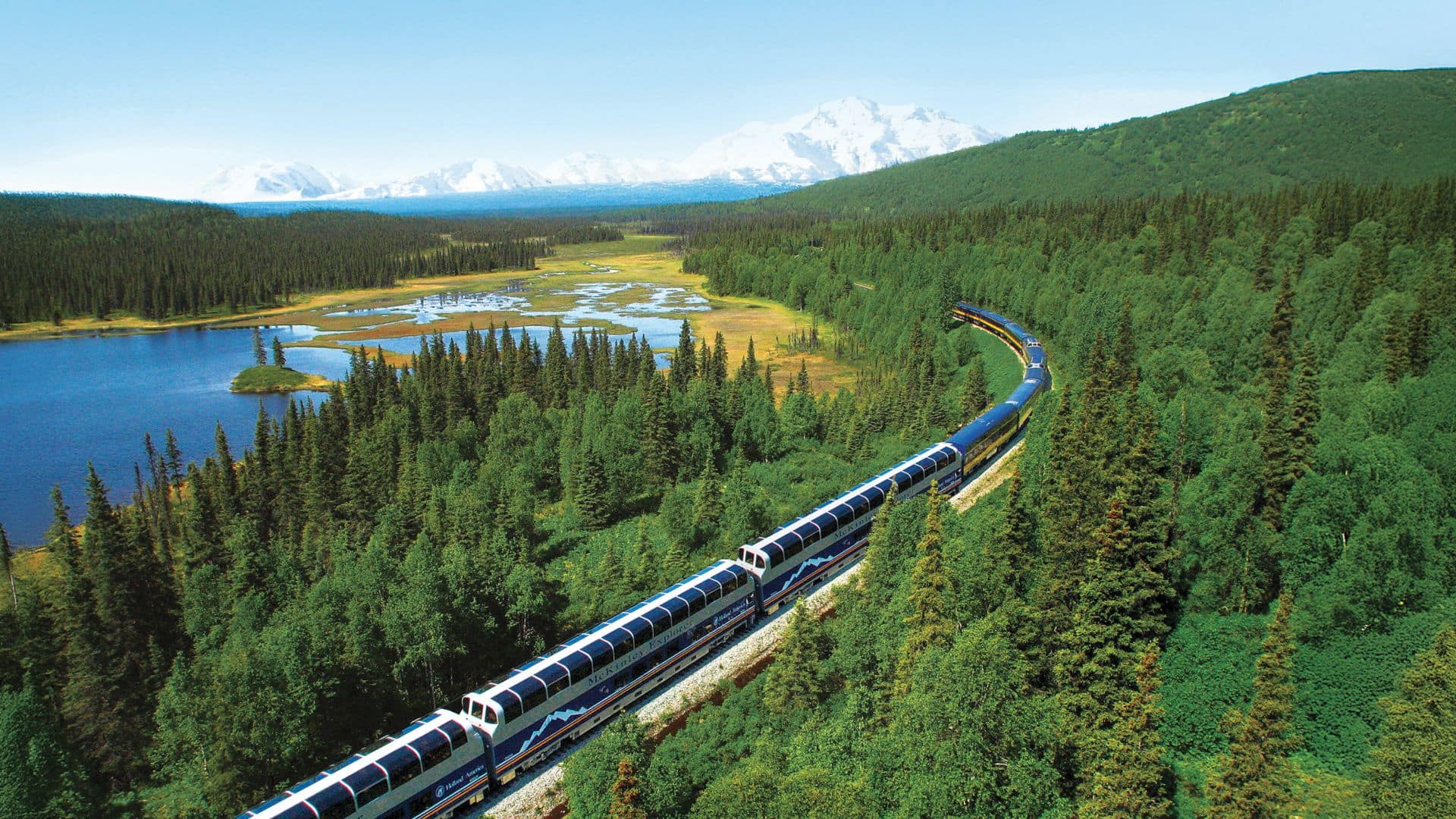 McKinley Explorer train traveling through a forested landscape with lakes and snow-capped mountains in the distance.