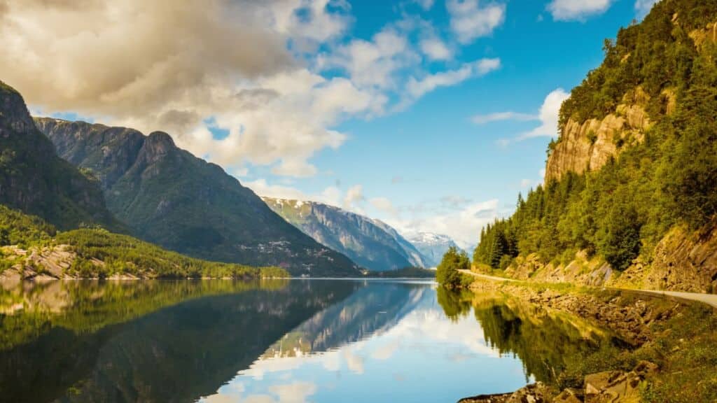 Mountain lake reflecting surrounding peaks and forested cliffs beside a winding shoreline road.