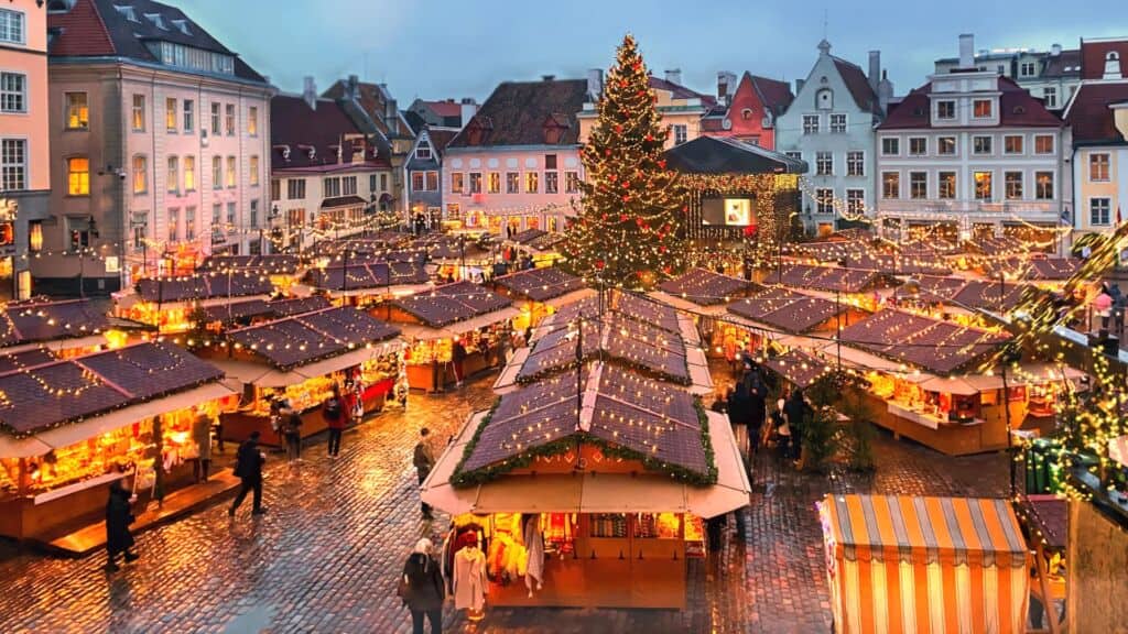 Festive European Christmas market at dusk, with glowing wooden stalls, a towering lit tree, cobblestone square, and historic pastel buildings among bundled shoppers.