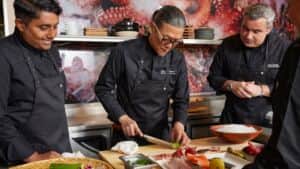 morimoto-guest-blog-header Chef Masaharu Morimoto slices a cucumber at a kitchen counter as two fellow chefs watch, surrounded by fresh fish, peppers, utensils and bowls beneath an octopus mural.