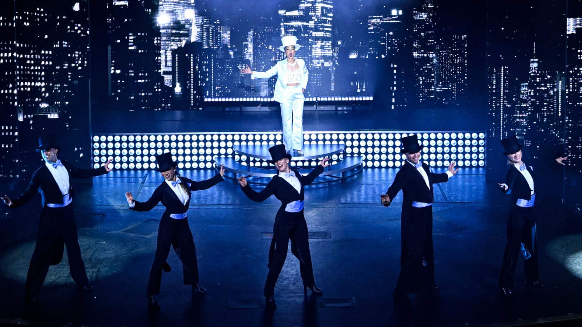 new-entertainment-shows-header-blog Five dancers in black tuxedos and top hats perform as a singer in white stands on a raised platform against a glittering city skyline.