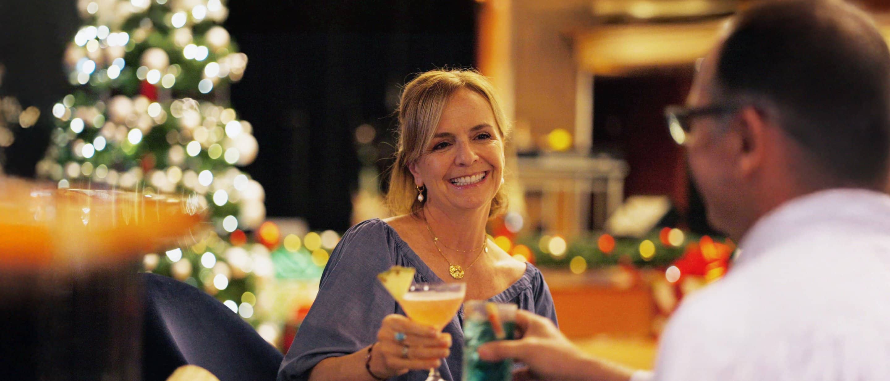 Two people enjoying drinks at a festive holiday gathering with a decorated Christmas tree in the background.