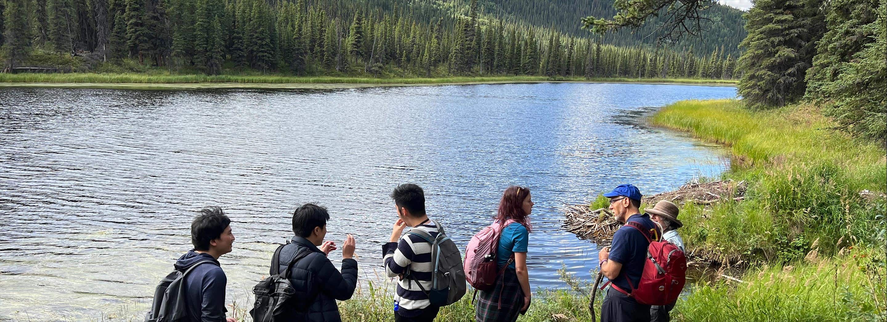 Group of hikers standing by a calm lake surrounded by evergreen forest and mountain scenery.