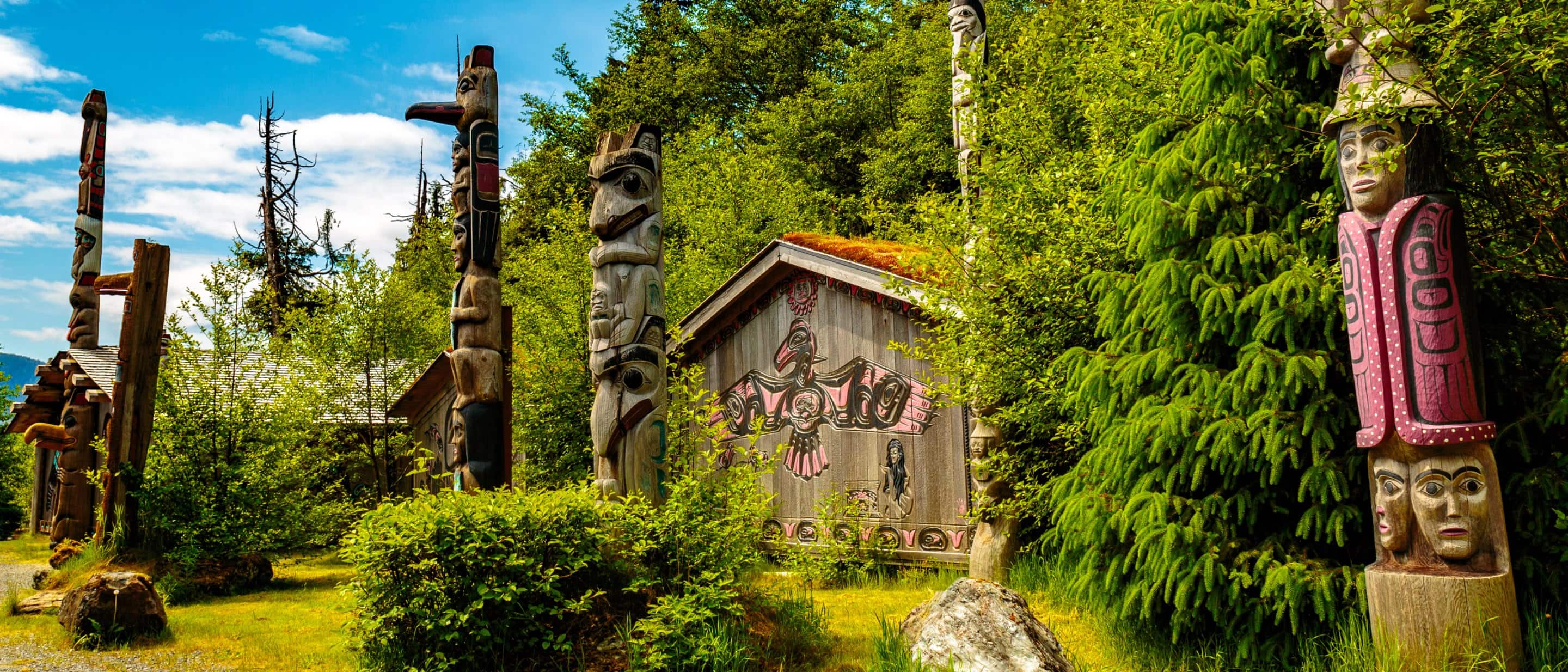 Colorful totem poles and traditional wooden clan house surrounded by lush green forest at Totem Bight State Park.