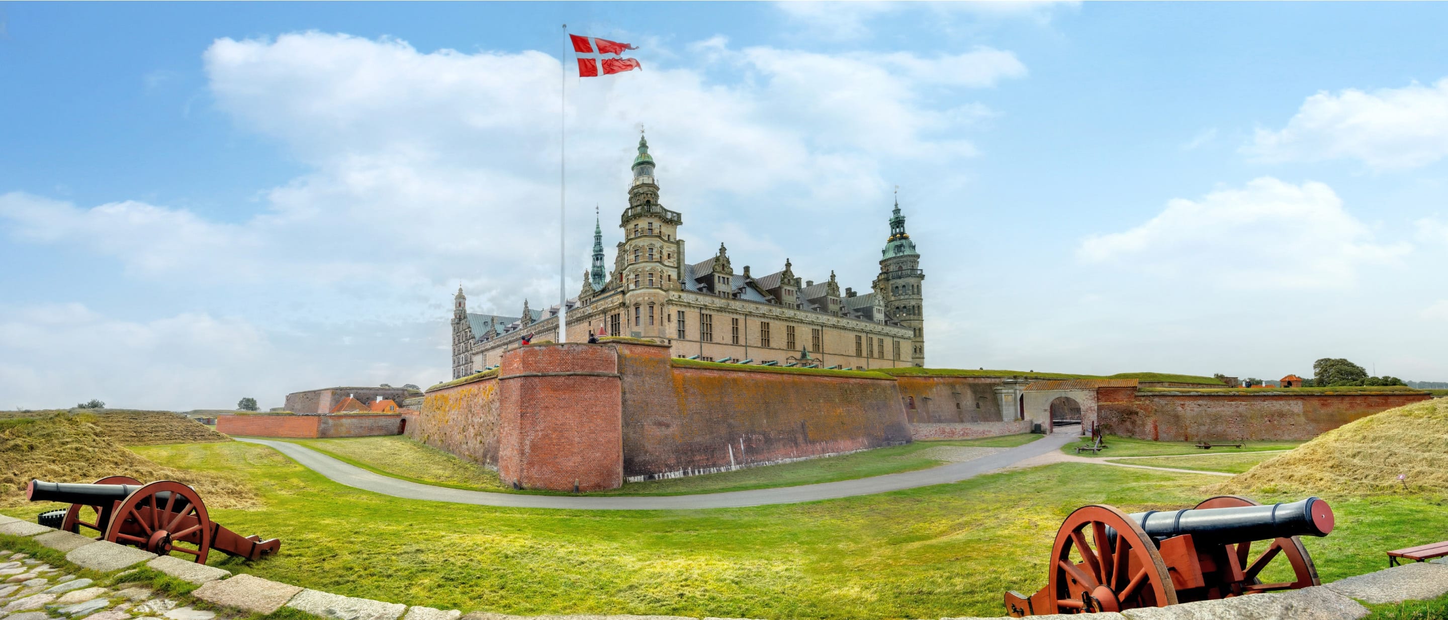 Historic Kronberg castle with Danish flag, brick walls, and cannons on grassy grounds near Copenhagen, Denmark.