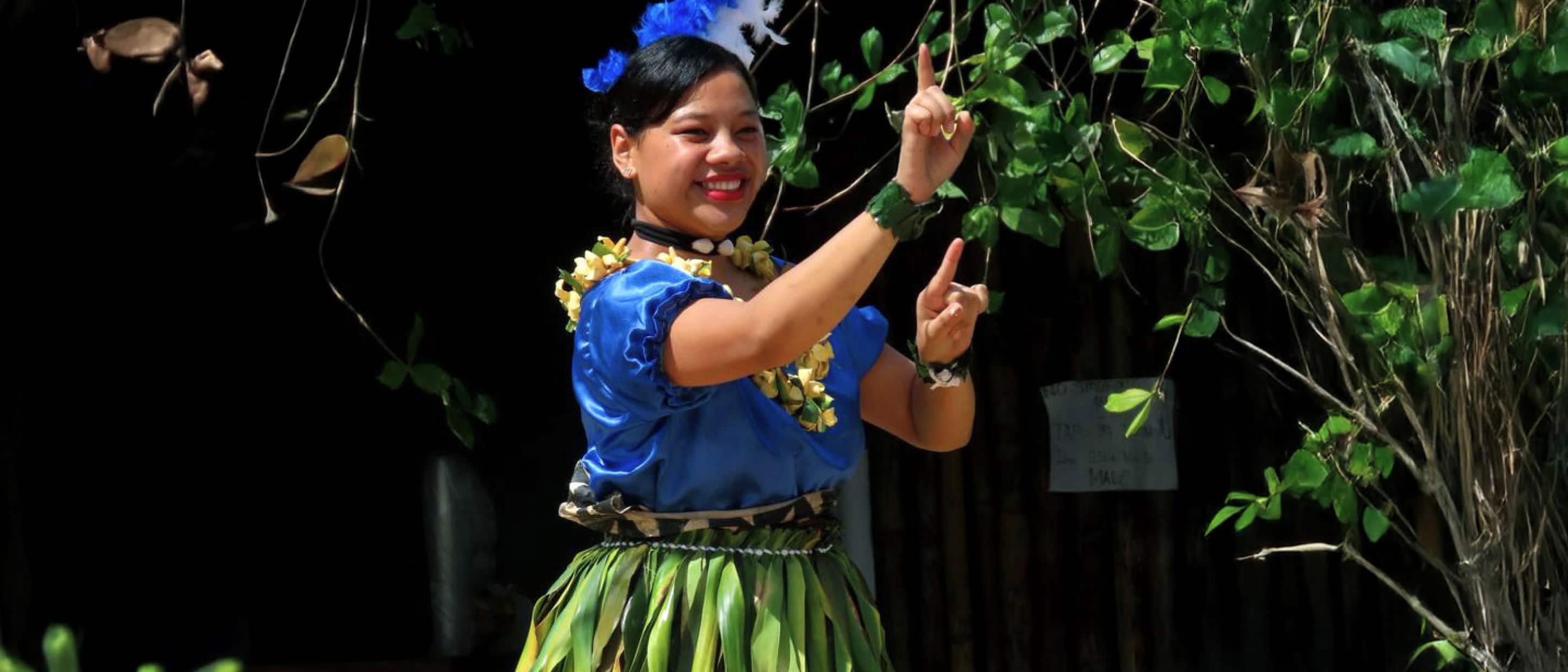 Traditional Polynesian dancer wearing blue top, green grass skirt, and floral garland performing cultural dance outdoors.