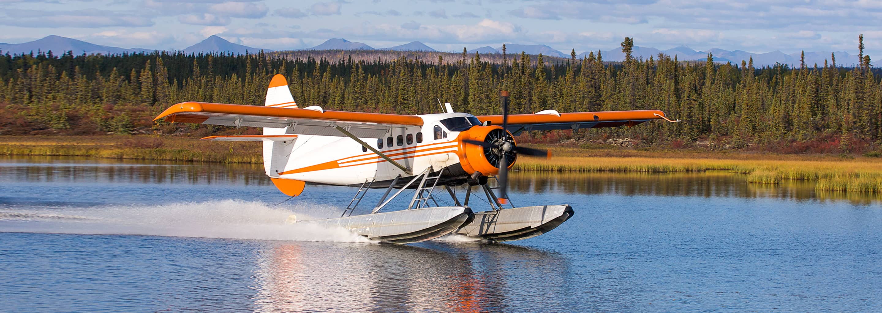 Orange and white floatplane seaplane taking off from an Alaskan lake with forest and mountains in the background.
