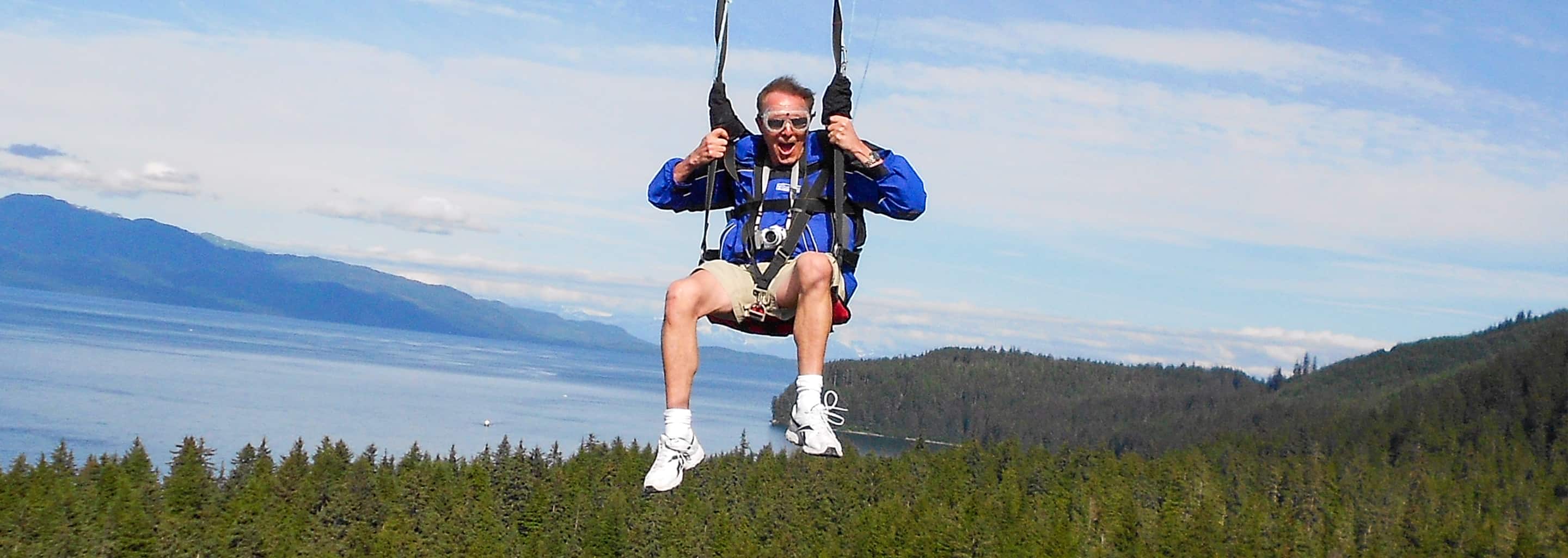 Man riding the world’s tallest zipline at Icy Strait Point in Alaska, soaring above a forest with ocean and mountains in the background.