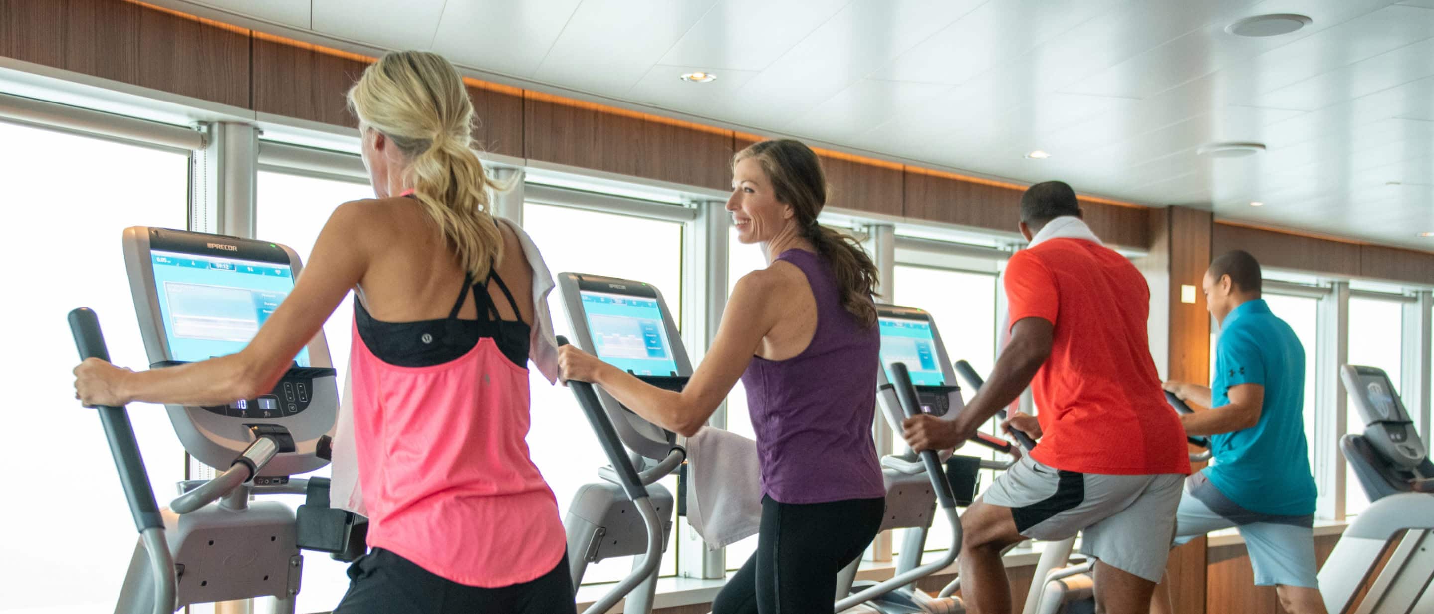 People using exercise machines in a fitness center on a Holland America cruise ship with large windows overlooking the water.