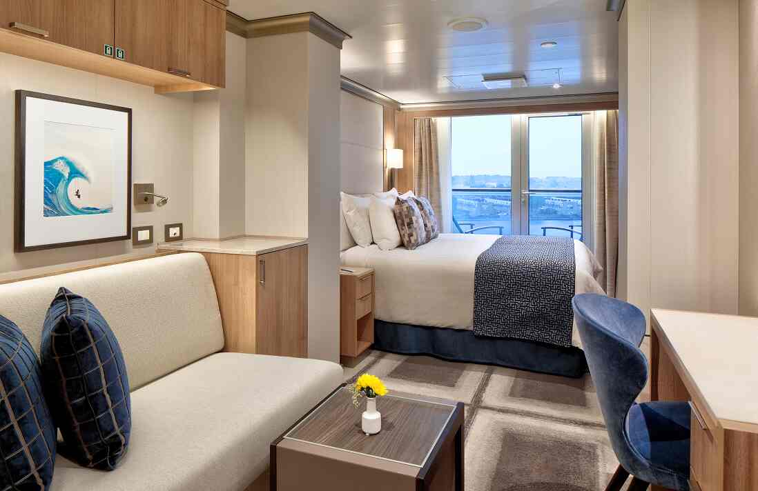 Aft View Vista stateroom with queen bed, sofa, coffee table, desk, wall art, and balcony on a Holland America cruise ship.