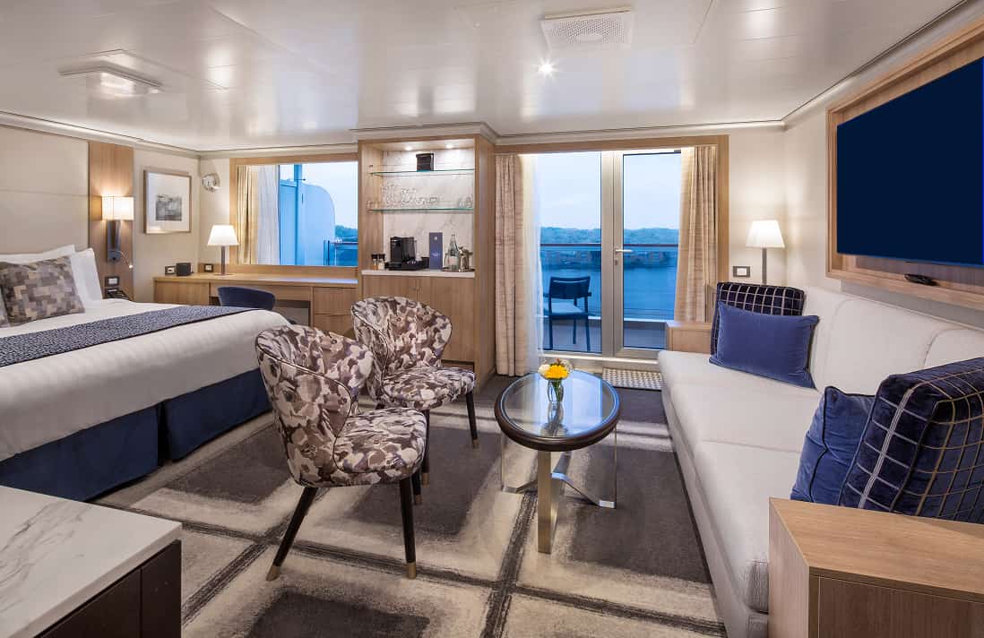 Neptune Suite Stateroom with king bed, patterned runner, two armchairs, sofa, coffee table, wall-mounted TV, and balcony on a Holland America cruise ship.