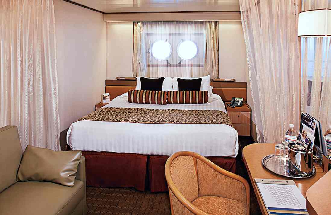 Porthole Ocean View stateroom with queen bed, chairs, and round window on Holland America cruise ship.