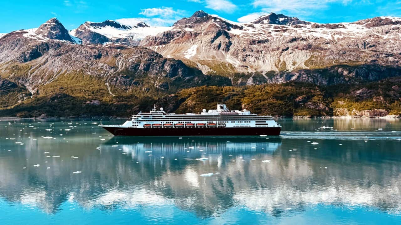 Holland America Line cruise ship sailing through icy Alaskan fjord with snow‑capped mountains.