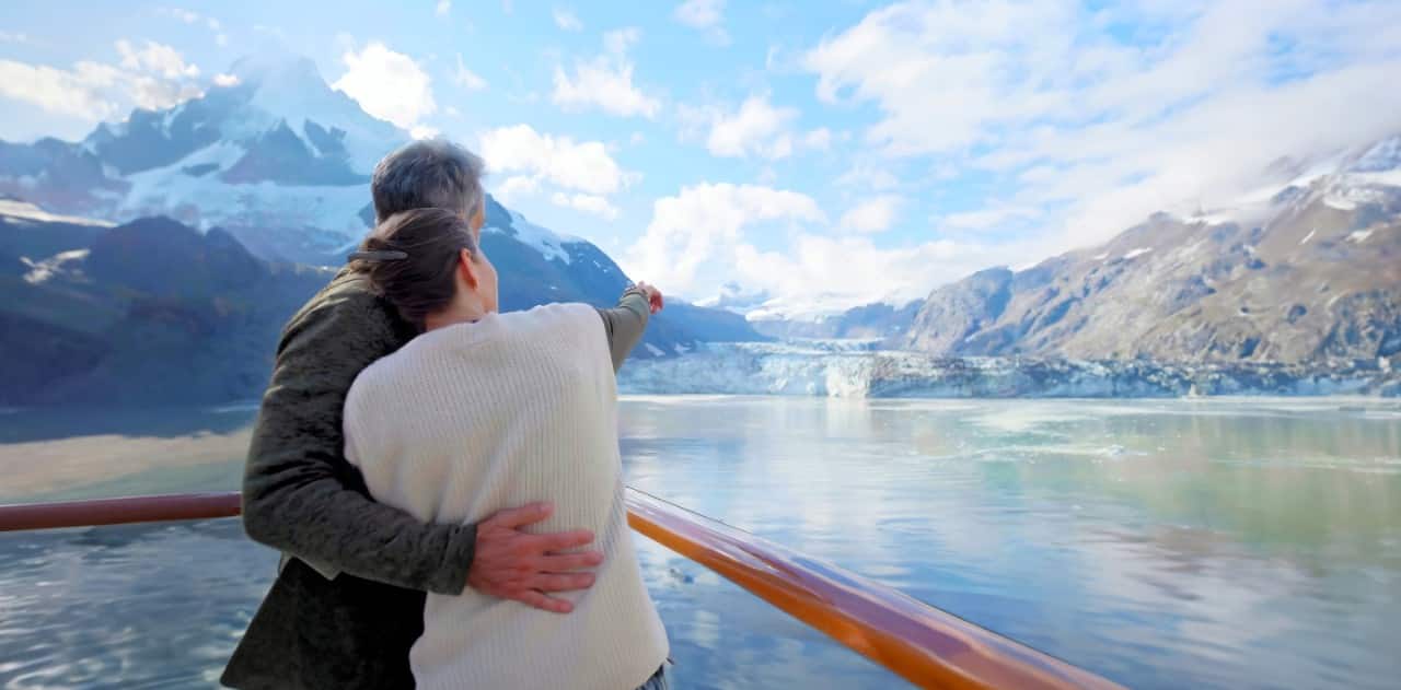 Couple standing on a ship’s viewing deck looking at a glacier surrounded by mountains under a bright blue sky.