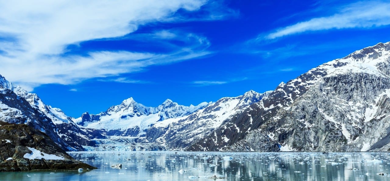 Glacier Bay with snow-covered mountains reflected in calm water with floating ice.