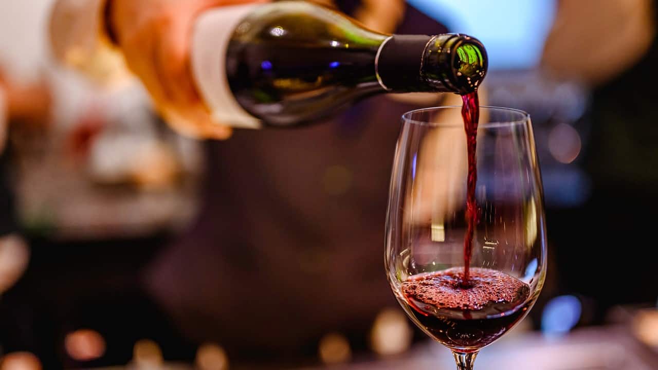 Close-up of red wine being poured from a bottle into a wine glass, with bubbles forming on the surface and a softly blurred restaurant or bar setting in the background.