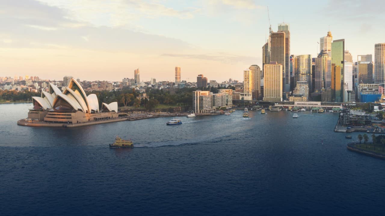 Sydney Harbour, Australia at sunset, featuring the iconic Sydney Opera House, city skyline, and boats on the water. 