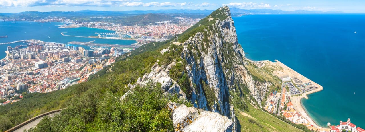 The Rock of Gibraltar rising steeply with cityscape and Mediterranean Sea in the background under a partly cloudy sky.