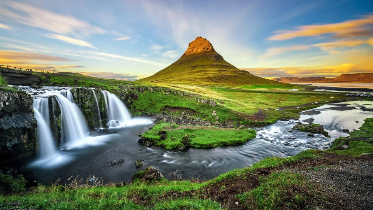 Pointed mountain, waterfall, and river in a lush Icelandic landscape at sunrise or sunset.