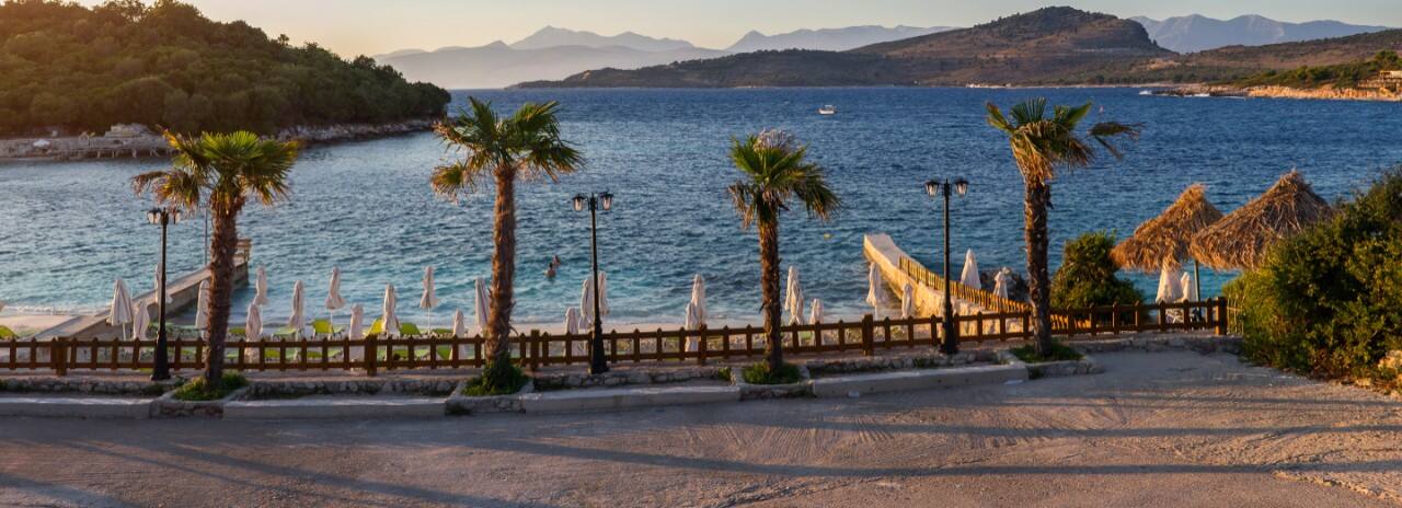 View of Palm Trees and a beach with umbrellas with water and mountains in the background.