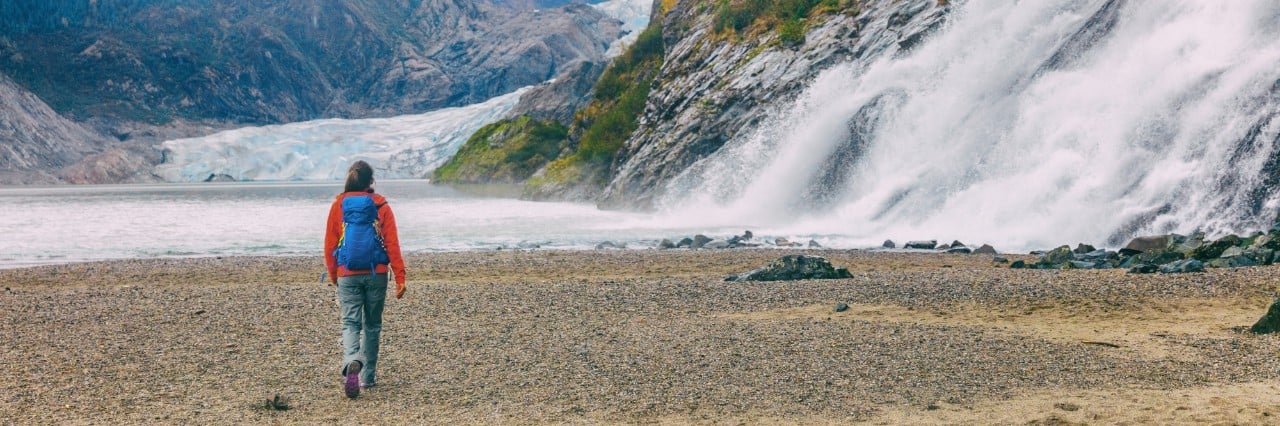 Hiker wearing a backpack walking along a rocky shoreline near a powerful waterfall with a glacier and rugged mountains in the background.