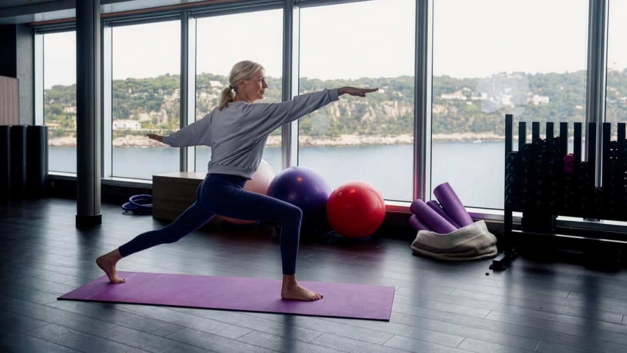 Person performing a yoga pose on a mat in a bright gym with large windows overlooking water