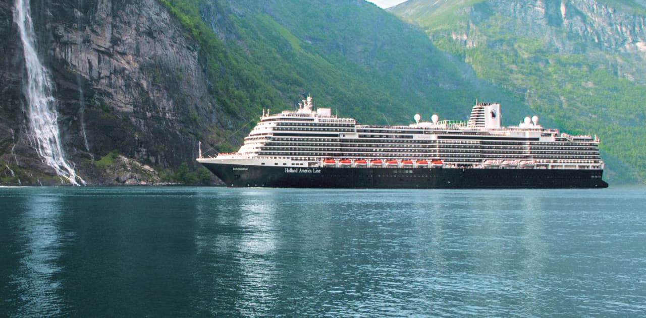 A Holland America Line ship sailing in front of green covered mountain and waterfall.