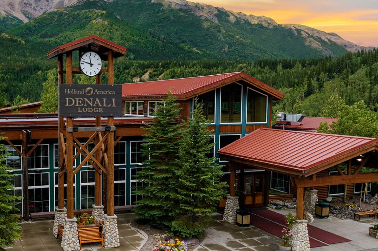 Holland America Denali Lodge with a rustic wooden clock tower, red metal roofs, and large glass windows, surrounded by lush greenery and mountains.