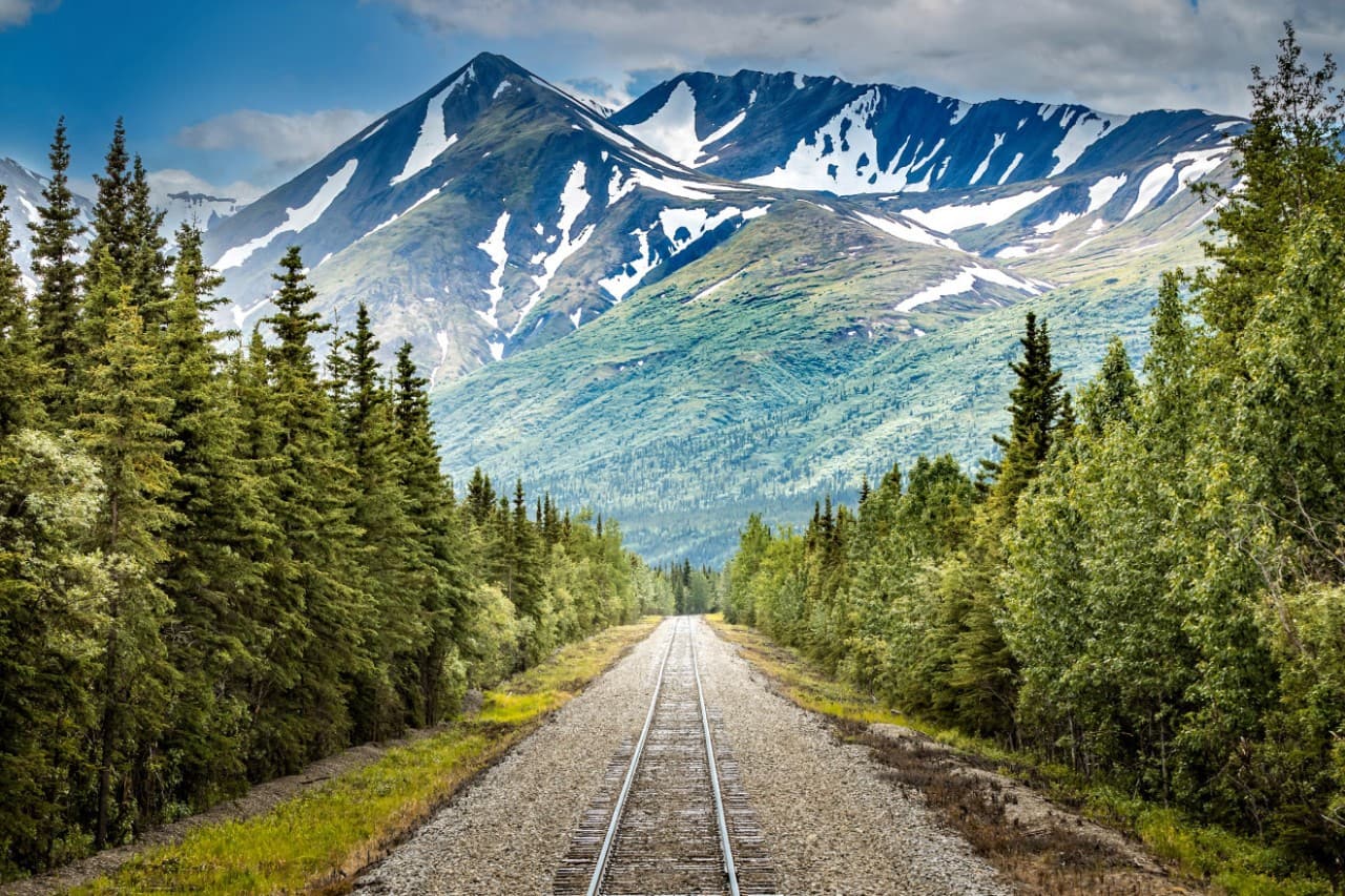 Holland America Denali Lodge with a rustic wooden clock tower, red metal roofs, and large glass windows, surrounded by lush greenery and mountains.