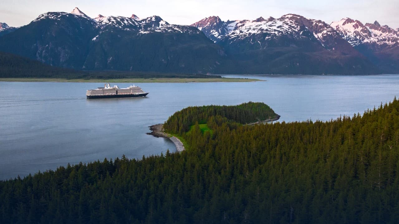 Panoramic view of a cruise ship navigating an Alaskan fjord, with forested islands and rugged snow-covered mountains in the background.