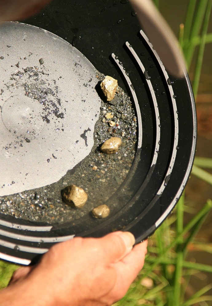 A close-up view of hands holding a black gold panning pan in water, with several gold nuggets visible among sand and gravel.