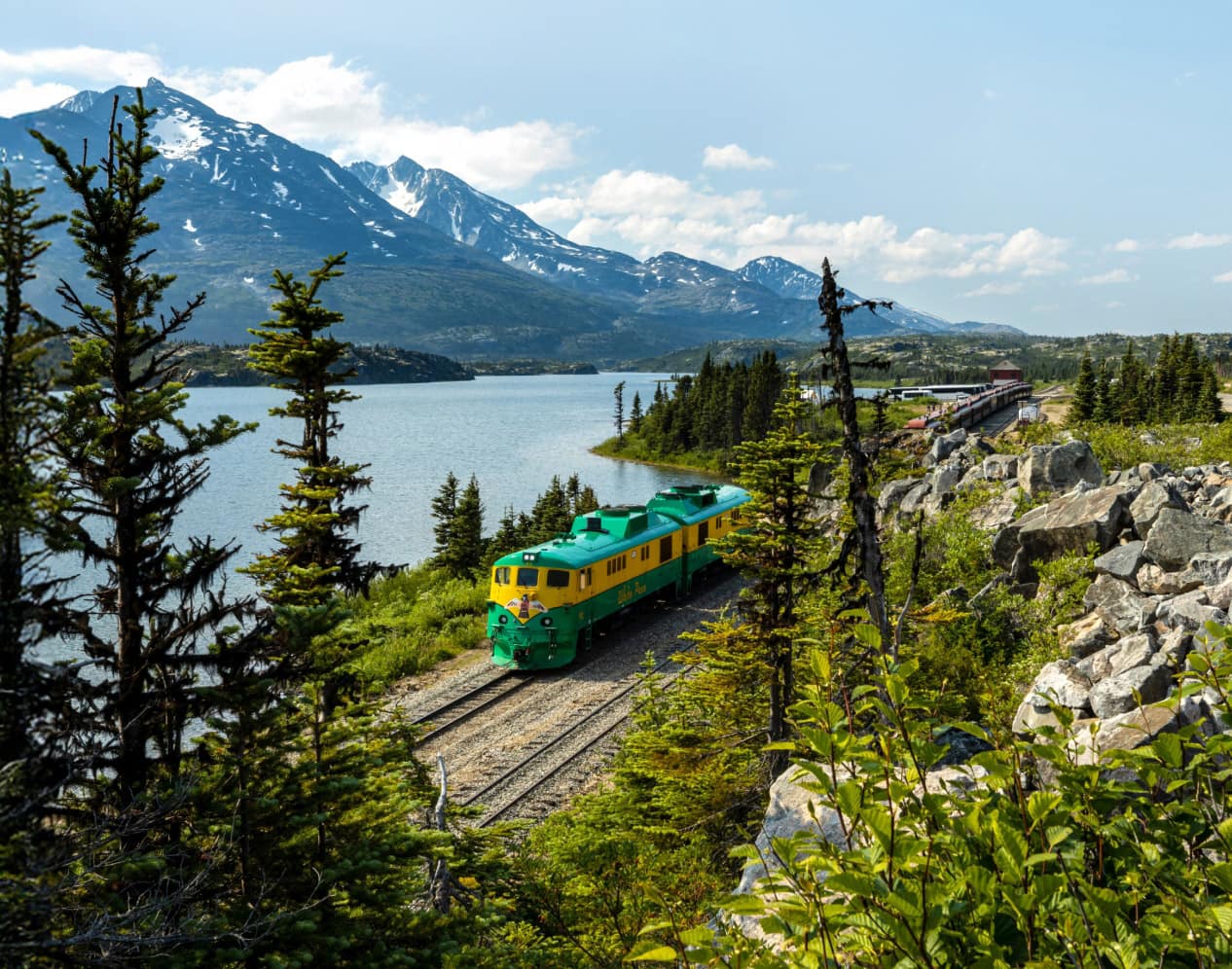 view of the White Pass & Yukon Route train, featuring a green and yellow locomotive on tracks alongside an Alaskan lake