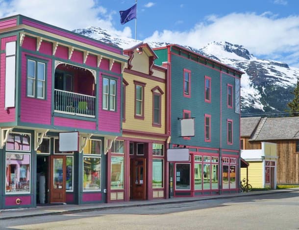 Colorful historic buildings line a street in downtown Skagway, Alaska, with snow-capped mountains in the background under a blue sky