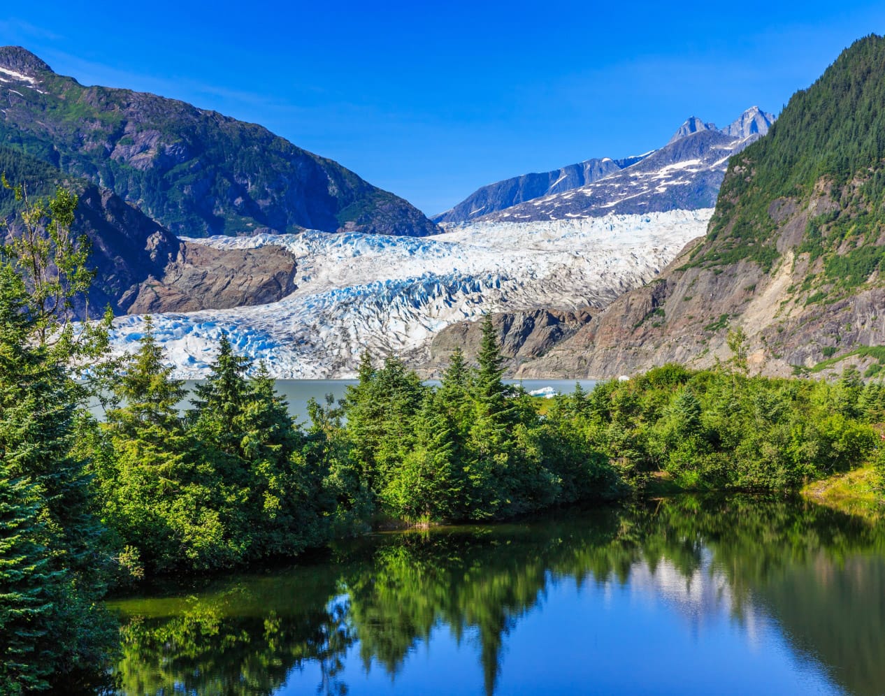 Mendenhall Glacier and surrounding mountains reflected in a calm lake, framed by green trees in the foreground.