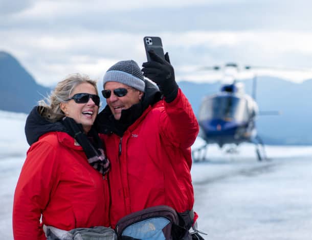Two smiling people in red jackets and sunglasses take a selfie on Mendenhall Glacier, with a helicopter in the background.