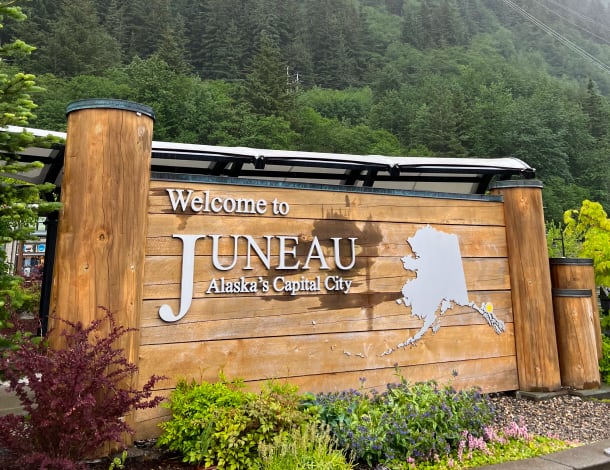 A wooden "Welcome to Juneau, Alaska's Capital City" sign with a map of Alaska, surrounded by greenery and backed by forested mountains.