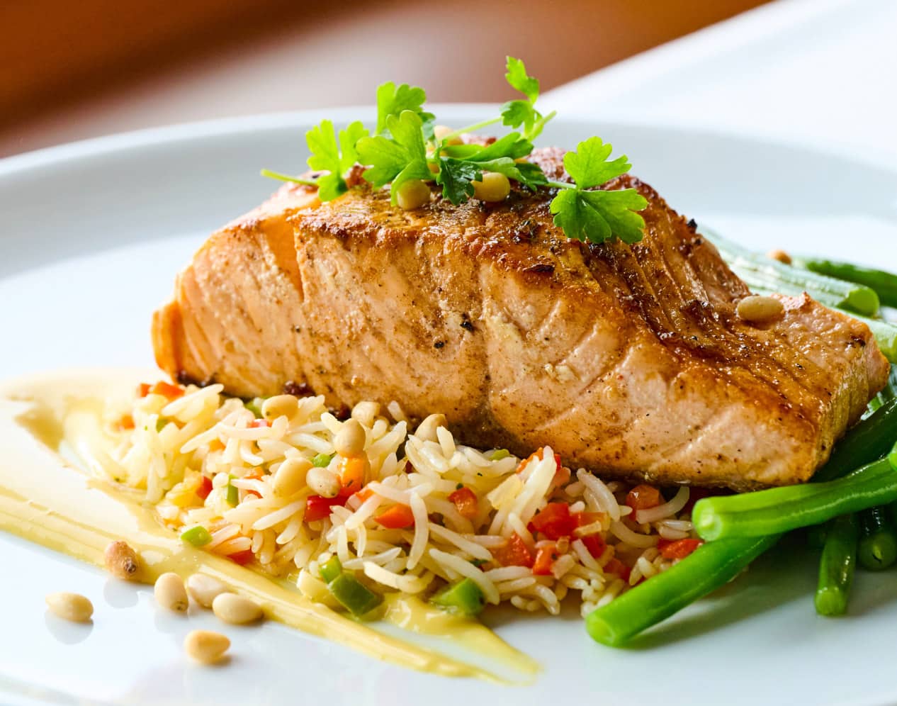 Pan-seared salmon fillet on a bed of rice pilaf with red peppers, served with green beans, garnished with fresh parsley and pine nuts