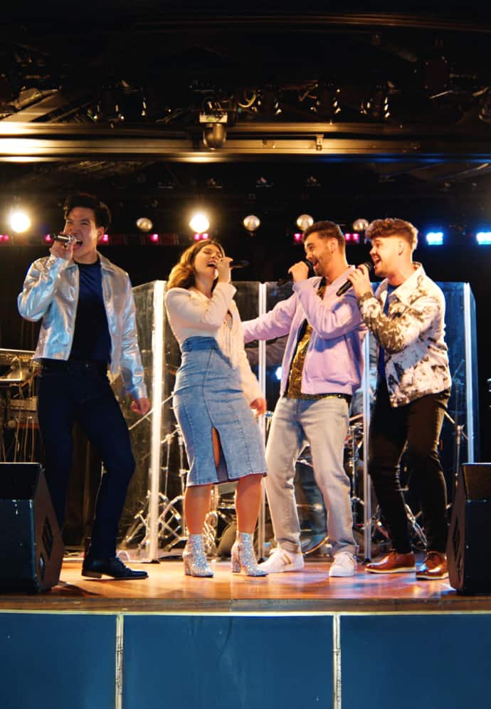 Four singers perform on stage with microphones at the Road House aboard a Holland America cruise ship