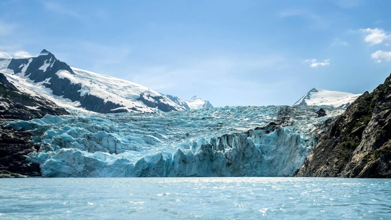Glacier flowing into a calm lake with snow covered mountains and rocky shores under a clear blue sky.