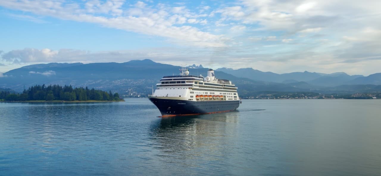 Holland America Cruise ship sails on calm water with mountains in the background.