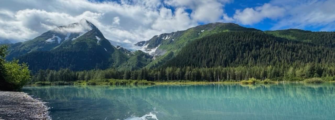 A kayak on turquoise water in Kenai Fjords National Park, Alaska, surrounded by forested mountains under a cloudy sky.