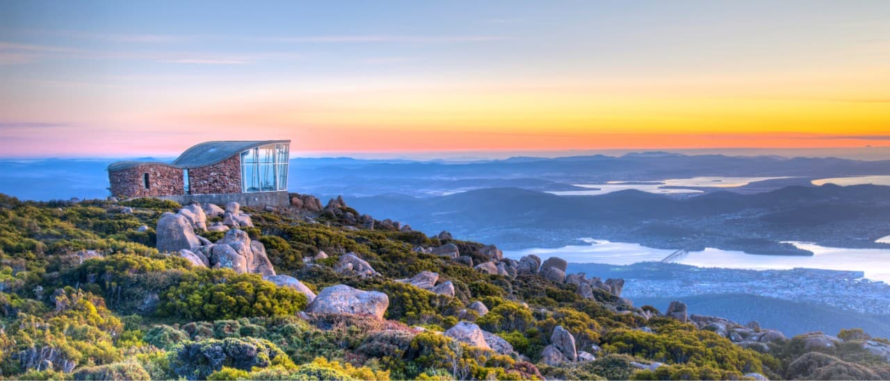 The top of Mount Wellington in Hobart, Tasmania, featuring a stone and glass lookout building, rugged terrain, and a sunset over the city and waterways below.