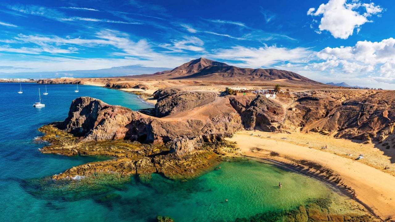 Rocky Canary Islands coastline with golden beaches, clear turquoise water, and volcanic hills under a blue sky.