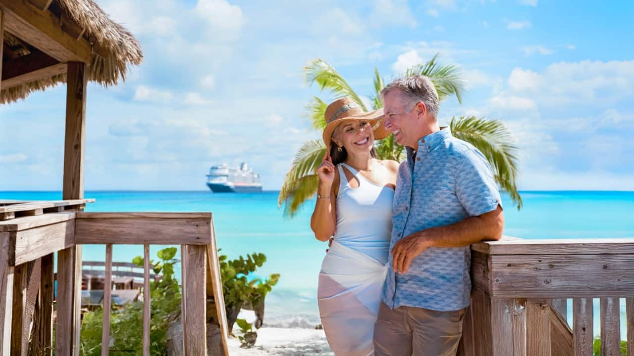 Two adults standing on a wooden beach deck with a thatched-roof hut, tropical plants, and turquoise ocean in the background, with a large cruise ship visible offshore.